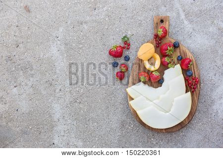 Top view on wooden board with fresh organic fruit and berries: melon strawberries blueberries apricot and red current on concrete background. Healthy snack