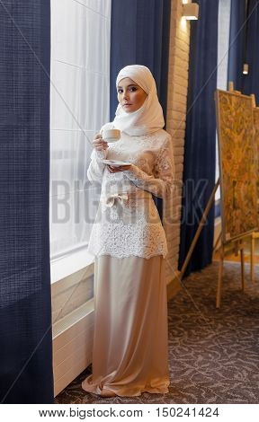 beautiful Muslim woman in a white wedding dress standing at window in hotel