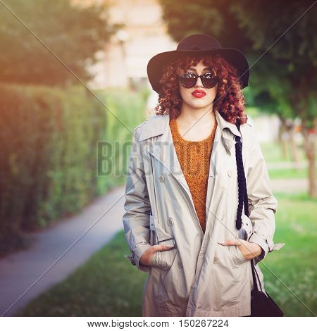 Modern stylish young woman in park in autumn. Closeup portrait of beautiful female person, in trench coat, floppy hat fall outfit standing in park. Retouched, filter applied, natural light.