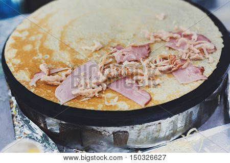 Making ham and cheese crepe at pan. Salty crepe or pancake with fillings made by street vendor's hands at outdoors creperie. French cuisine, cooking for commercial kitchen.