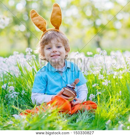Cute happy little boy wearing Easter bunny ears at spring green grass and blooming apple garden, eating chocolate muffing with bunny and having fun outdoors.