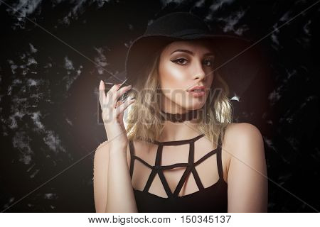 Closeup studio portrait of gorgeous blonde young lady in black dress and floppy hat with makeup, on black background. Retouched, back lit.