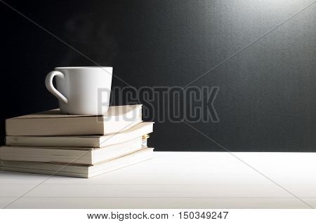 Still life - A cup of hot coffee put on old books on white wooden table in font of grunge black background.