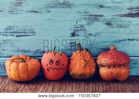 an apple disguised as a pumpkin with a funny face trying to go unnoticed between different real pumpkins, on a rustic wooden surface, against a blue wooden background