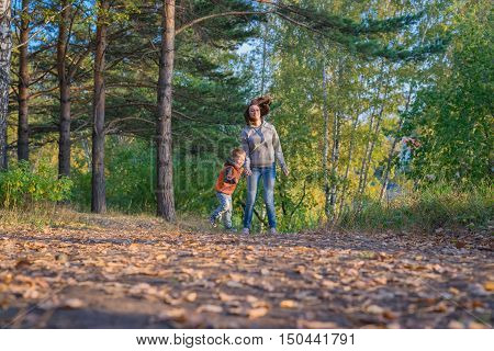 Mother and son running on path in autumn forest. Family at nature autumn bright sunny day fallen yellow leaves underfoot.