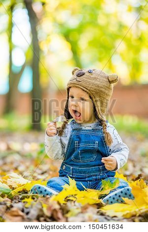 Happy happy baby sitting in the park with yellow leaves in autumn in bear cap. In autumn park kid sitting in yellow leaves. Cheerful baby sitting in the park in bear cap. Warm autumn day in the park.