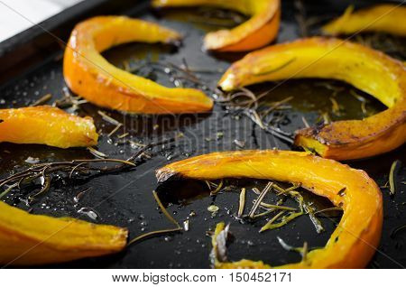 Freshly baked and juicy slices of pumpkin with addition aromatic herbs and olive oil on a baking tray. Pumpkin slices arranged on rows. Thanksgiving Halloween holidays concept. Selective focus.