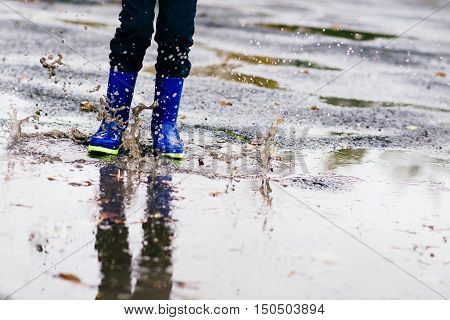 Boy In Rubber Blue Rainboots Jumping To Dirty Puddle