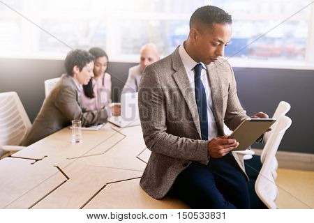 Businessman sitting on the edge of the board room table holding and using an electronic tablet with three colleagues seated at the table behind him.