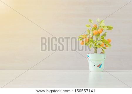 Closeup artificial orange plant with orange fruit in green pot on blurred wooden desk and wall textured background in office room for decorate under window light with copy space