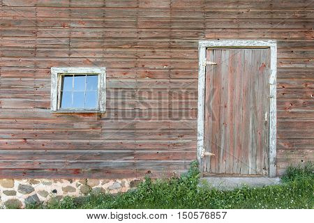 Section of a weathered building showing a door and single pane window. Textured wood with faded red grey and white framing.