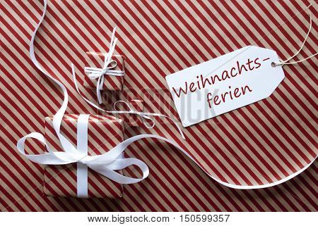 Two Gifts Or Presents With White Ribbon. Red And Brown Striped Wrapping Paper. Christmas Or Greeting Card. Label With German Text Weihnachtsferien Means Christmas Break