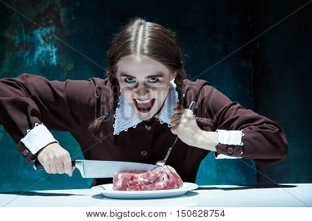 Portrait of a young girl with knife, fork and and a piece of raw meat on a plate. Girl in school uniform as killer. The image in the style of Halloween and Addams family