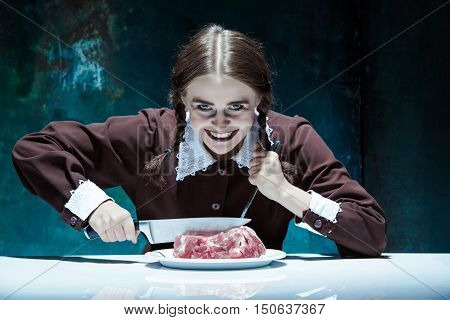 Portrait of a young girl with knife fork and and a piece of raw meat on a plate. Girl in school uniform as killer. The image in the style of Halloween and Addams family