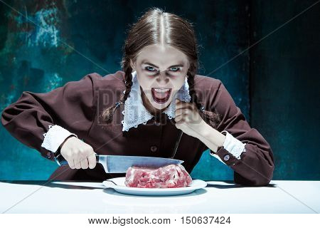 Portrait of a young girl with knife fork and and a piece of raw meat on a plate. Girl in school uniform as killer. The image in the style of Halloween and Addams family