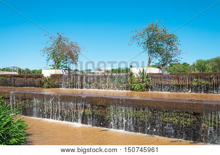Falling cascading water of a lake in a park. Vegetation around with plants and some flowers. Beautiful sunny day with blue sky background. Natural wonders of South America.