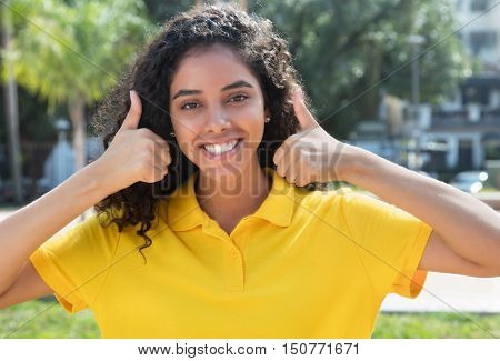 Beautiful latin american girl with long dark hair showing both thumbs outdoors in the summer in the city