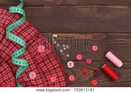 Red fabric , thread, measuring tape and buttons on the wooden table. The view from the top. Sewing accessories.