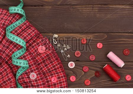 Red fabric , thread, measuring tape and buttons on the wooden table. The view from the top. Sewing accessories.