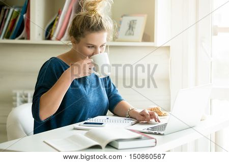 Portrait of young beautiful smiling casual woman sitting at office desk enjoying her cup of coffee while working or studying on laptop computer at small home office or in the student dorm. Indoors