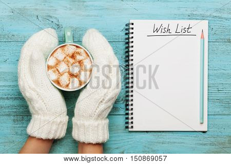 Woman hands in mittens hold cup of hot cocoa or chocolate with marshmallow and notebook with wish list on turquoise vintage table from above, christmas planning concept. Flat lay style.
