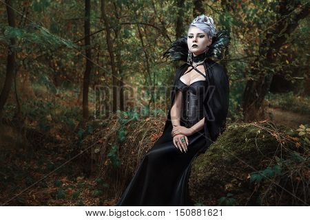 Gothic girl in black raincoat sits on a tree in the forest.