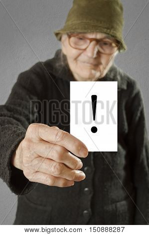 Elderly woman holding card with printed exclamation mark. Selective focus on card and fingers.