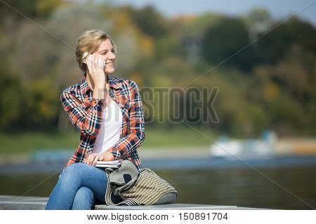 Portrait of a young attrractive girl sitting her legs crossed, head turned, at the bridge, talking on her phone, waterline behind, lifestyle, copyspace
