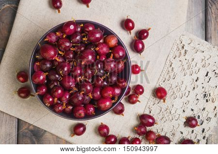Ripe red purple berry gooseberries in purple bowl on gray wooden table. Gooseberry berries on the table cloth. Top view.