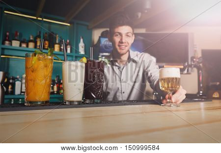 Young handsome Barman offers exotic sweet non-alcoholic cocktails in restaurant. Professional male bartender at work in bar made refreshing drinks. Selective focus on glass of cream soda