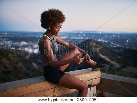 athletic african american woman using smartphone at runyon canyon while resting on bench at dusk