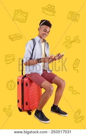 Using A Card. Positive Relaxed Teenager Sitting On His Big Suitcase And Smiling While Holding A Smar