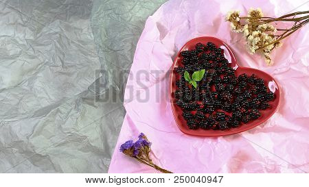 Black Raspberry On A Decorative Red Plate In The Form Of A Heart, With Dry Flowers On The Pepper Bac