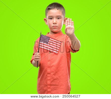 Dark haired little child holding United States flag with open hand doing stop sign with serious and confident expression, defense gesture