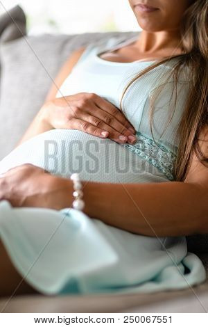 Close-up Of Pregnant Woman In Dress Relaxing And Sitting On The Side On The Sofa. Holding A Hands On