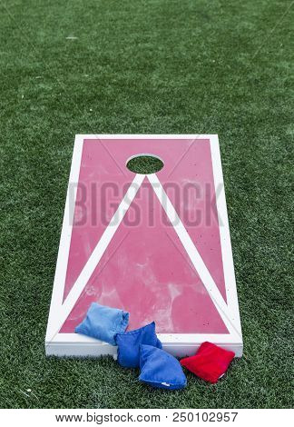 A Wood Red And White Corn Hole Board Is Set Up On A Green Turf Field With Blue And Red Bean Bags.