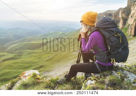 Smiling Girl Traveler In A Yellow Hat And A Pair Of Sunglasses Ssitting At The Foot Of Epic Rocks Wi
