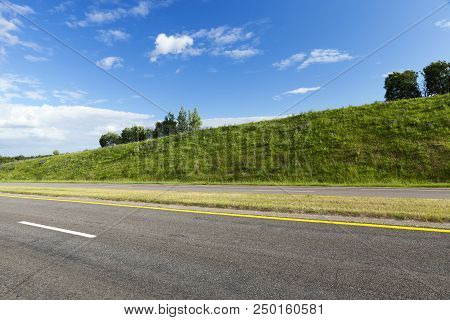 Wide Asphalt Road Divided Into Several Lanes, Around The Road There Are Hills With Green Grass, Summ