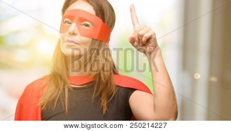 Middle age super hero woman wearing red mask and cape raising finger, the number one