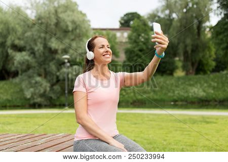 fitness, technology and healthy lifestyle concept - smiling woman in headphones listening to music and taking selfie by smartphone at park