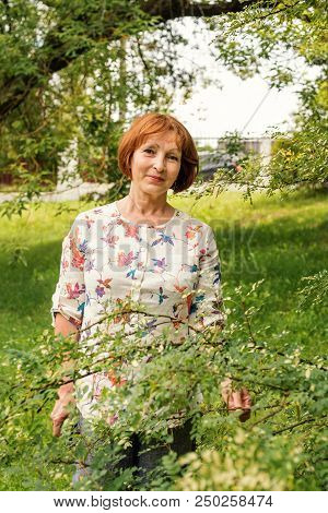 A Beautiful Elderly Woman In Flaxen Clothes Stands Tranquilly In Her Green Garden