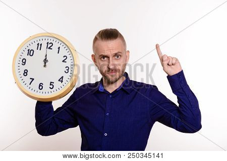 Strict Man Holding A Large Wall Clock And Points A Finger On Empty Space On White Background