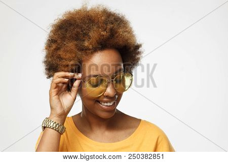 Shy Dark Skin Afrcian Female Model Looking Down, Holding Her Yellow Eyeglasses, Isolated On Gray Bac