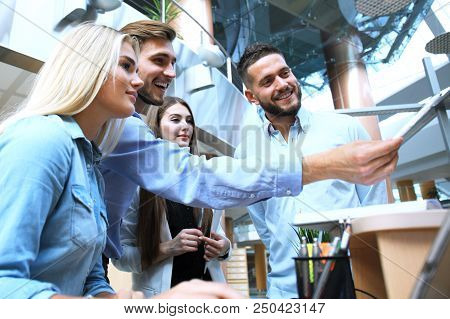 Group Of Young People In Casual Wear Sitting At The Office Desk And Discussing Something While Looki
