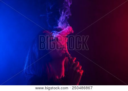 Young Woman In Neon Light And Smoke Of E-cigarettes Or Vape On Dark Background.
