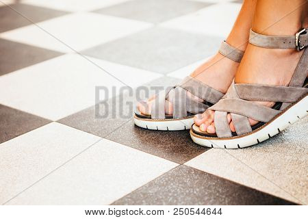 Female Feet With A Nude Pedicure In Beige Suede Sandals On The Background Of A Chessboard Floor.
