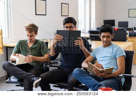 Student Hiding Behind Textbook And Sitting Between Two Friends. Three Fellow Students Studying In Cl