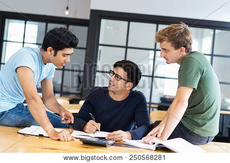 Three Multiethnic Fellow Students Doing Homework Together. Young Men Sitting On Desk And Discussing 