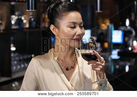 Beautiful Relaxed Asian Businesswoman Standing At Bar Counter Smelling Glass Of Wine With Closed Eye