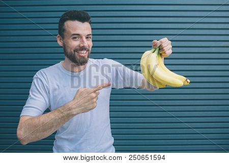Happy Guy Is Hlding Bananas In One Hand And Pointing On Them With Another One. He Is Looking On Came
